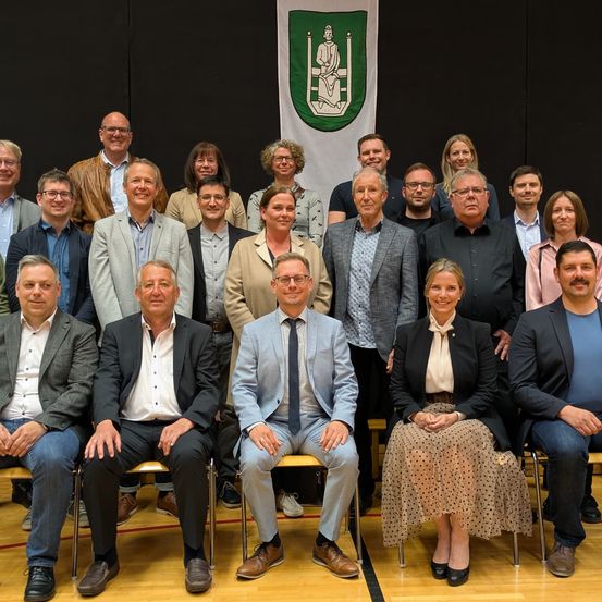 Eine Gruppe von Personen, darunter Männer und Frauen, in formeller Kleidung, versammelt sich zu einem Foto auf einem Holzboden. Hinter ihnen befindet sich ein grüner Banner mit einem Logo.