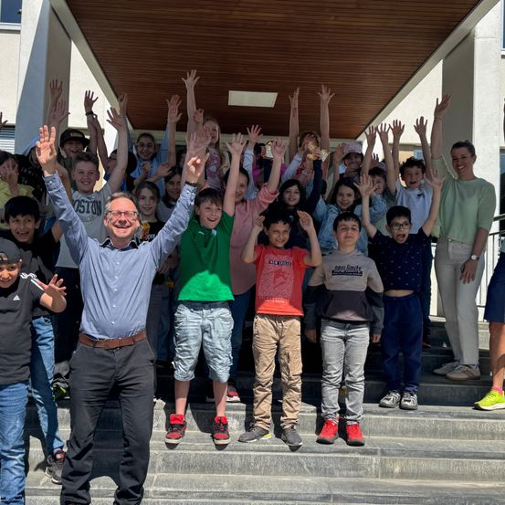 Eine Gruppe von Kindern und Erwachsenen steht auf den Stufen und hebt die Hände. Ein Mann in einem blauen Hemd und schwarzen Hosen steht vor ihnen.