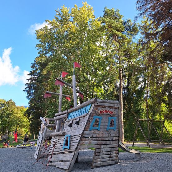Ein hölzerne Piratenschiff-Spielplatz mit roten Flaggen oben in einem Park mit Kiesboden. Rechts befindet sich eine Rutsche. Es gibt Bäume und Menschen im Hintergrund.