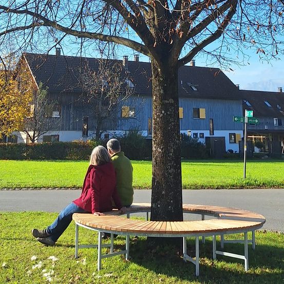 Zwei Menschen sitzen auf einer geschwungenen Bank unter einem Baum und sehen sich an. Sie sind in einem Park mit einem Gebäude im Hintergrund.