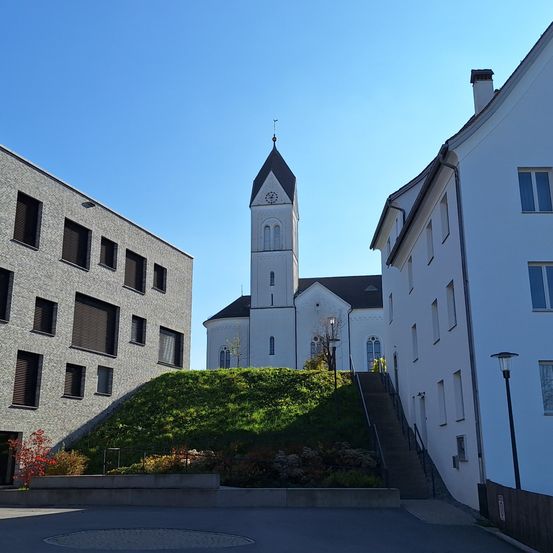Eine Kirche mit einem hohen Turm und einer Uhr steht einem modernen Gebäude mit Fenstern gegenüber, vor dem sich ein Garten unter einem klaren blauen Himmel befindet.