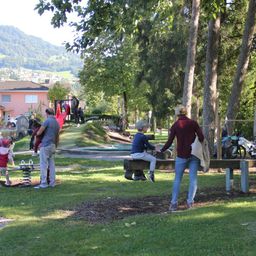 Eine Gruppe von Menschen, darunter Kinder, befindet sich in einem Park. Ein Kind rutscht eine Rutsche hinunter, während andere auf einer Bank sitzen. Eine Frau geht in der Nähe.