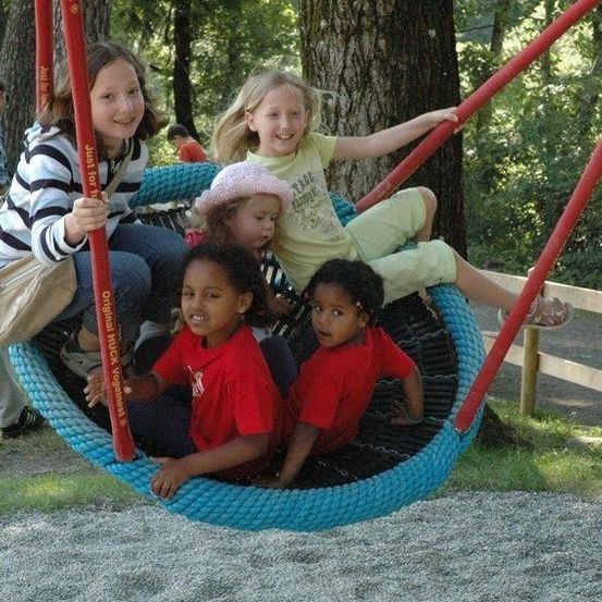 Vier Kinder in roten T-Shirts schwingen sich in einem blauen Reifenschaukel, während ein Mädchen in einem gelben T-Shirt sich festhält. Eine Frau in einem gestreiften T-Shirt geht vorbei.