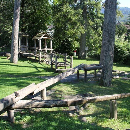 Ein Rasenbereich mit Holzstrukturen, einem Baumstamm und einem Pilz. Die Holzstrukturen umfassen eine Bank und ein kleines Haus. Bäume und Pflanzen sind in der Umgebung.