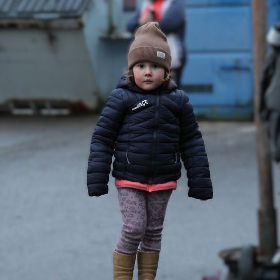 Ein kleines Mädchen in einer marineblauen Jacke, einem rosa Shirt, gemusterten Leggings und braunen Stiefeln steht auf einem Bürgersteig mit einer blauen Wand und einer Person im Hintergrund.