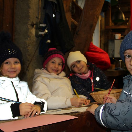 Drei Kinder sitzen an einem Holzschreibtisch, lächeln und halten Stifte. Ein Kind trägt einen blauen Hut, ein anderes einen rosa Hut und das dritte einen weißen Hut. Ein Korb und Jacken sind im Hintergrund.
