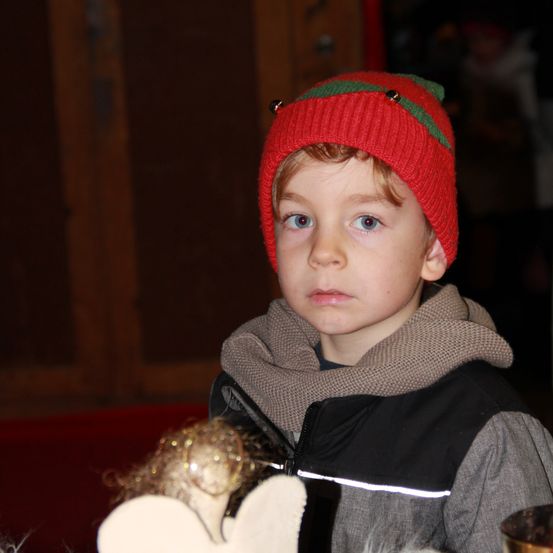 Ein junger Junge mit roten Haaren trägt eine rote Mütze mit grünem Rand, eine graue Jacke und einen braunen Schal. Er hält eine goldene Engelsfigur in der Hand.
