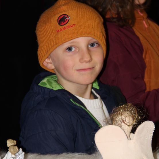 Ein Junge mit einer orangefarbenen Mütze und einem lächelnden Gesicht sitzt und hält eine kleine Engelsfigur. Er trägt eine blaue Jacke mit grünem Reißverschluss.