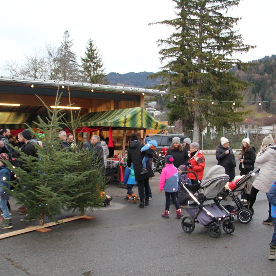 Ein Weihnachtsbaummarkt mit Menschen, die sich um einen Baum versammelt haben, einige in Winterkleidung. Ein Kinderwagen und ein Auto sind in der Nähe geparkt.