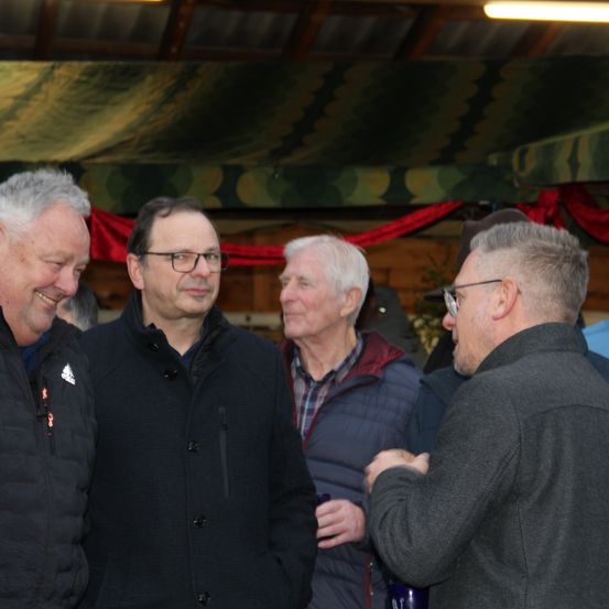 Vier Männer in Jacken stehen in einem Raum. Ein Mann trägt eine Brille und hält ein Getränk. Ein anderer lächelt, und die anderen scheinen in ein Gespräch vertieft zu sein.