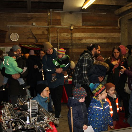 Eine Gruppe von Erwachsenen und Kindern versammelt sich drinnen, einige halten Säuglinge. Links ist ein großes mechanisches Objekt ausgestellt, während andere sich unterhalten oder beobachten. Der Raum hat Holzwände und eine Uhr an der Wand.