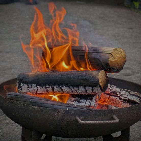 Eine Nahaufnahme eines Feuerkorbs mit brennenden Holzscheiten. Die Flammen sind hell orange und der Rauch steigt auf. Der Feuerkorb ist aus Metall und hat einen Griff an der Seite.