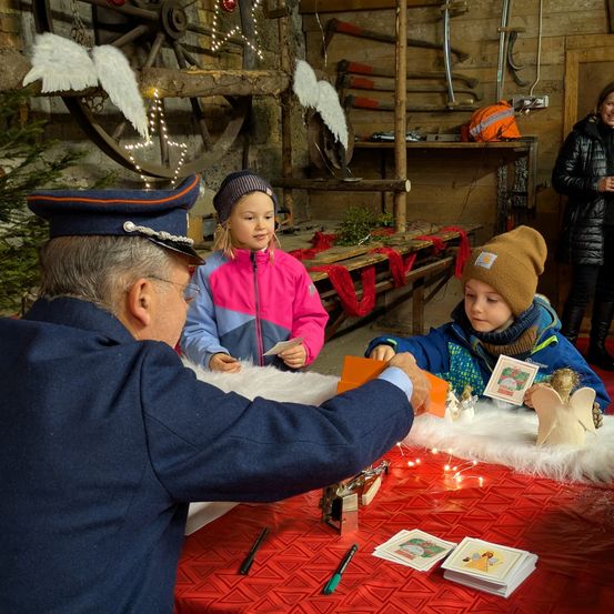 Ein älterer Mann in Uniform hilft zwei Kindern an einem festlich geschmückten Weihnachtstisch, der mit Karten und Engeldekorationen verziert ist.