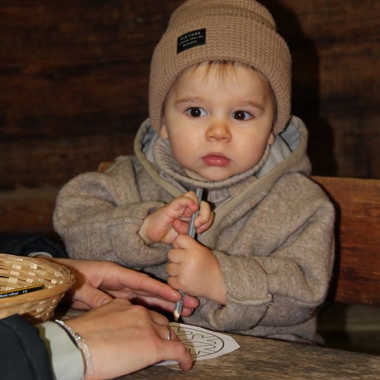 Ein Baby sitzt an einem Tisch und hält einen Bleistift. Es trägt eine braune Mütze und eine Jacke. Eine andere Person hält den Stift und führt die Hand des Babys.