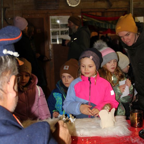 Eine Gruppe von Kindern in Winterkleidung steht um einen Tisch herum und beobachtet, wie ein Erwachsener einen Tonengel macht.