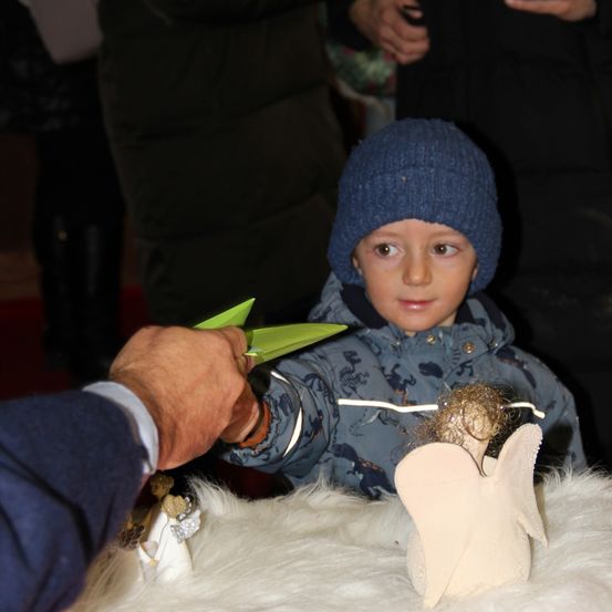 Ein Kind in einer blauen Mütze und Jacke hält ein Papierflugzeug und schaut auf eine weiße Engelsfigur auf einem weißen Pelztuch.