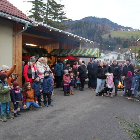 Eine Menschenmenge, darunter Familien und Kinder, versammelt sich um einen festlichen Bereich mit Ständen und einem Lagerfeuer. Die Umgebung ist im Freien, mit Bergen und Häusern im Hintergrund.