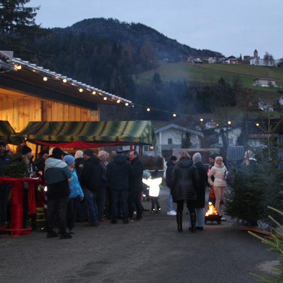 Eine Gruppe von Menschen steht um einen Weihnachtsbaummarkt, einige mit Mützen und Winterkleidung. Eine Person steht vor einem Feuer.