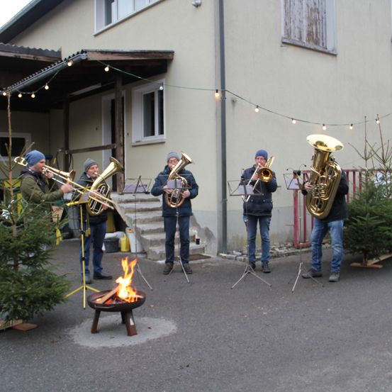 Fünf Männer spielen Tubas vor einem Gebäude mit einem Feuerkorb davor. Über ihnen hängen Lichterketten.