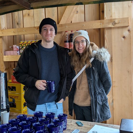 Ein Mann und eine Frau stehen hinter einem Tisch mit blauen Tassen und lächeln für ein Foto. Sie befinden sich in einem Raum mit Holzwänden und Regalen voller Gegenstände.