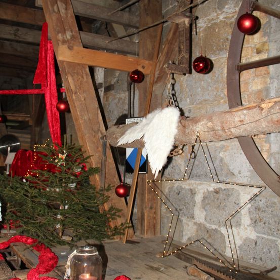 Ein rustikaler Innenbereich mit einem Weihnachtsbaum, einem hölzernen Engel und einer Sterndekoration. Der Raum hat Holzbalken, Ziegelwände und rote Ornamente. Eine Kerze steht auf einem Glas auf einem Holztisch.