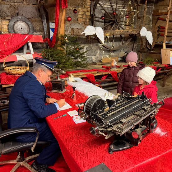 Ein Mann in Uniform sitzt an einem Tisch mit einer Schreibmaschine und schreibt Briefe. Zwei Kinder in Winterkleidung stehen neben ihm und lächeln. Die Umgebung ist mit einem Weihnachtsbaum und Dekorationen für Weihnachten geschmückt.