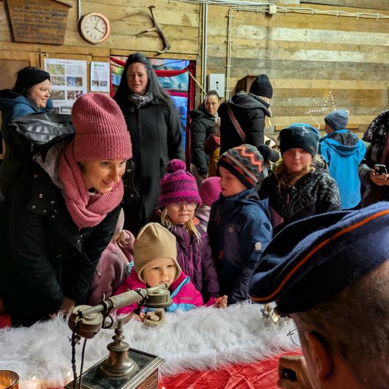 Eine Gruppe von Menschen, darunter Frauen und Kinder, versammelt sich um ein altes Telefon auf einem mit Fell bedeckten Tisch. Sie sind warm für das kalte Wetter gekleidet. Hinter ihnen zeigt eine Holzwand eine Uhr und Poster.