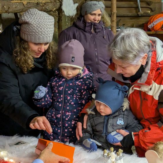 Drei Frauen und zwei Kinder öffnen ein Geschenk an einem Tisch, wobei ein Mann es präsentiert. Sie tragen Winterkleidung, wobei ein Kind eine Blumenjacke und ein anderes eine blaue Mütze trägt.