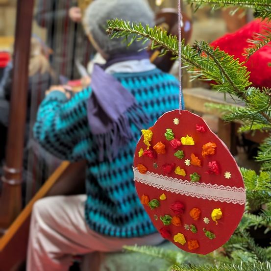 Eine Person in einem blauen Pullover und einer lila Schürze sitzt auf einer Bank und spielt eine Harfe, während ein festlicher roter Weihnachtsschmuck von einem Weihnachtsbaum hängt.