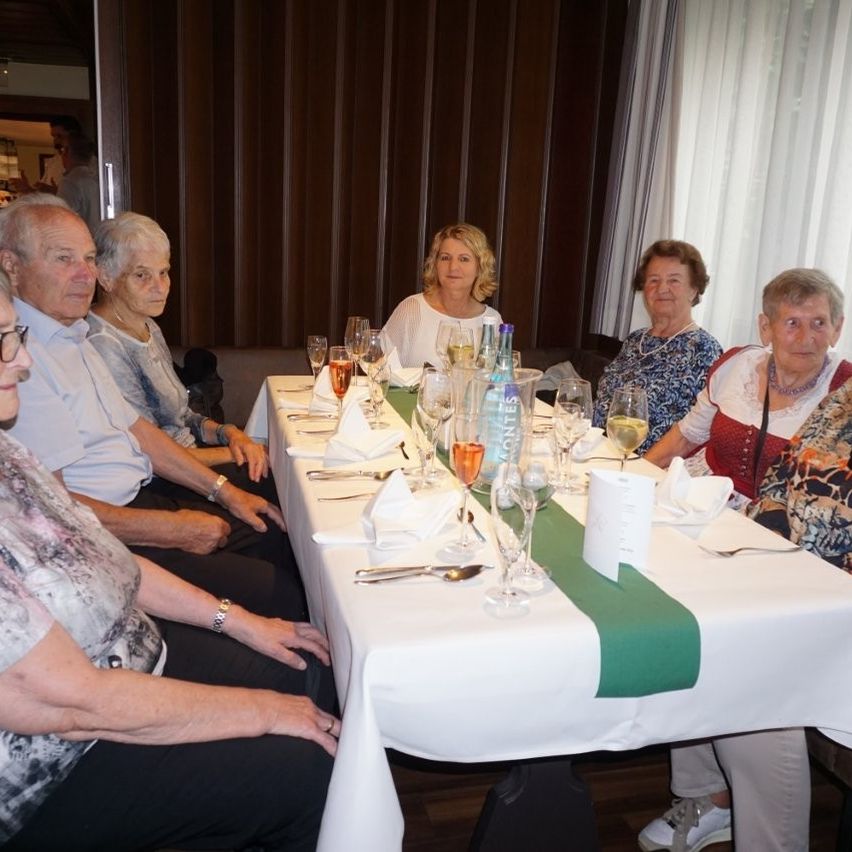 Eine Gruppe älterer Menschen sitzt um einen Tisch, der für eine Mahlzeit gedeckt ist, trägt Freizeitkleidung und Brillen. Der Tisch ist mit einer weißen Tischdecke bedeckt und hat Weingläser, eine Flasche und Servietten. Hinter ihnen führt eine Tür in einen anderen Raum.