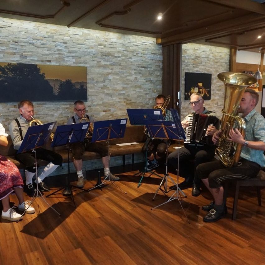 Eine Gruppe von Musikern spielt Instrumente in einem Raum. Sie sitzen auf einer Bank und stehen auf einem Holzboden. Einige von ihnen tragen Brillen und Lederschuhe.