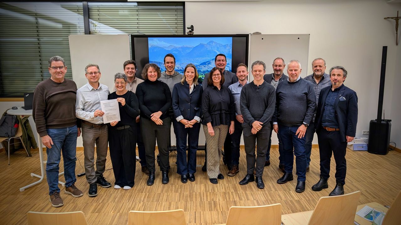 Eine Gruppe von Menschen steht in einem Raum mit einem großen Bildschirm, auf dem Berge im Hintergrund zu sehen sind. Sie scheinen für ein Foto zu posieren, lächeln und schauen in die Kamera.