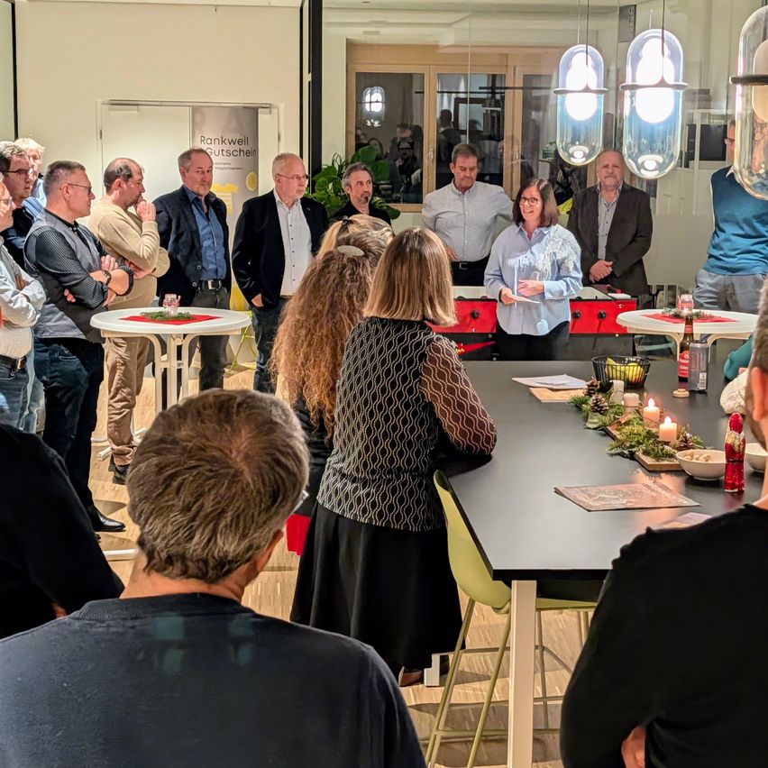 Eine Gruppe Erwachsener versammelt sich in einem Raum mit einem Tisch, auf dem einige Kerzen und dekorative Gegenstände stehen. Eine Frau steht am Tisch, während andere aufmerksam zuhören.