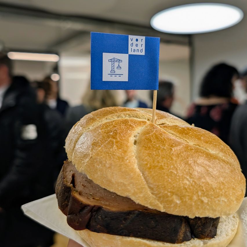 Ein Sandwich mit einer blauen Flagge mit der Aufschrift 'Vor der Land' auf einem Zahnstocher, gehalten in einer Hand mit Menschen im Hintergrund.