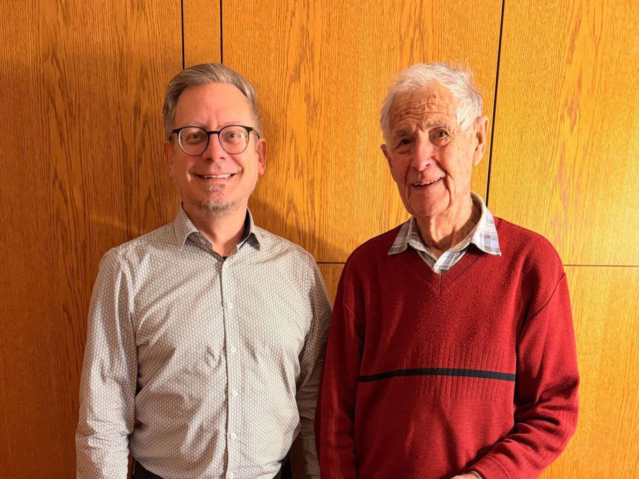 Zwei Männer, einer mit einer gemusterten Bluse und einer Brille, der andere in einem roten Pullover, stehen eng nebeneinander an einer Holzwand und lächeln beide.