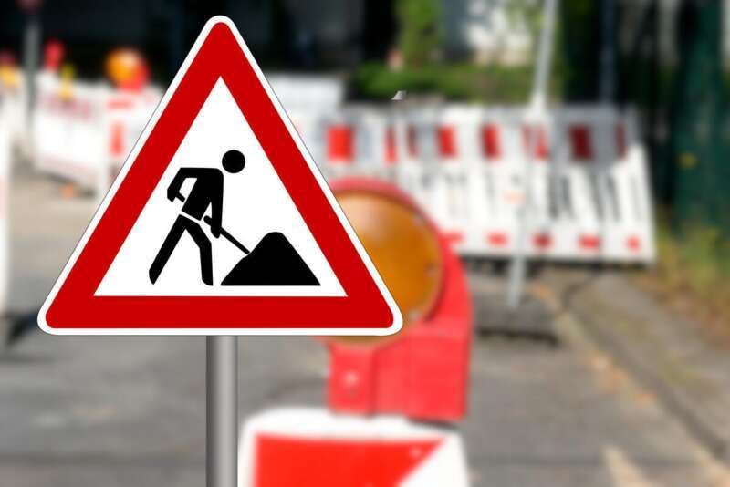 A road sign with a figure digging is on a post near traffic cones. There is a blurry view of a road with plants.
