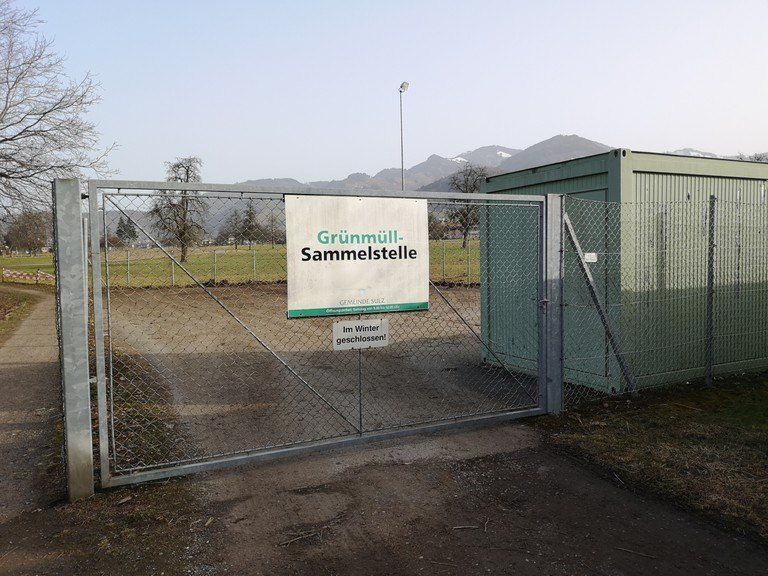 Ein geschlossenes Tor mit einem Schild, das auf eine Grünabfallsammelstelle hinweist. Das Tor ist von einem Metallzaun umgeben. Hinter dem Tor steht ein grüner Container. Im Hintergrund befindet sich eine weite Wiese mit Bäumen und Bergen.