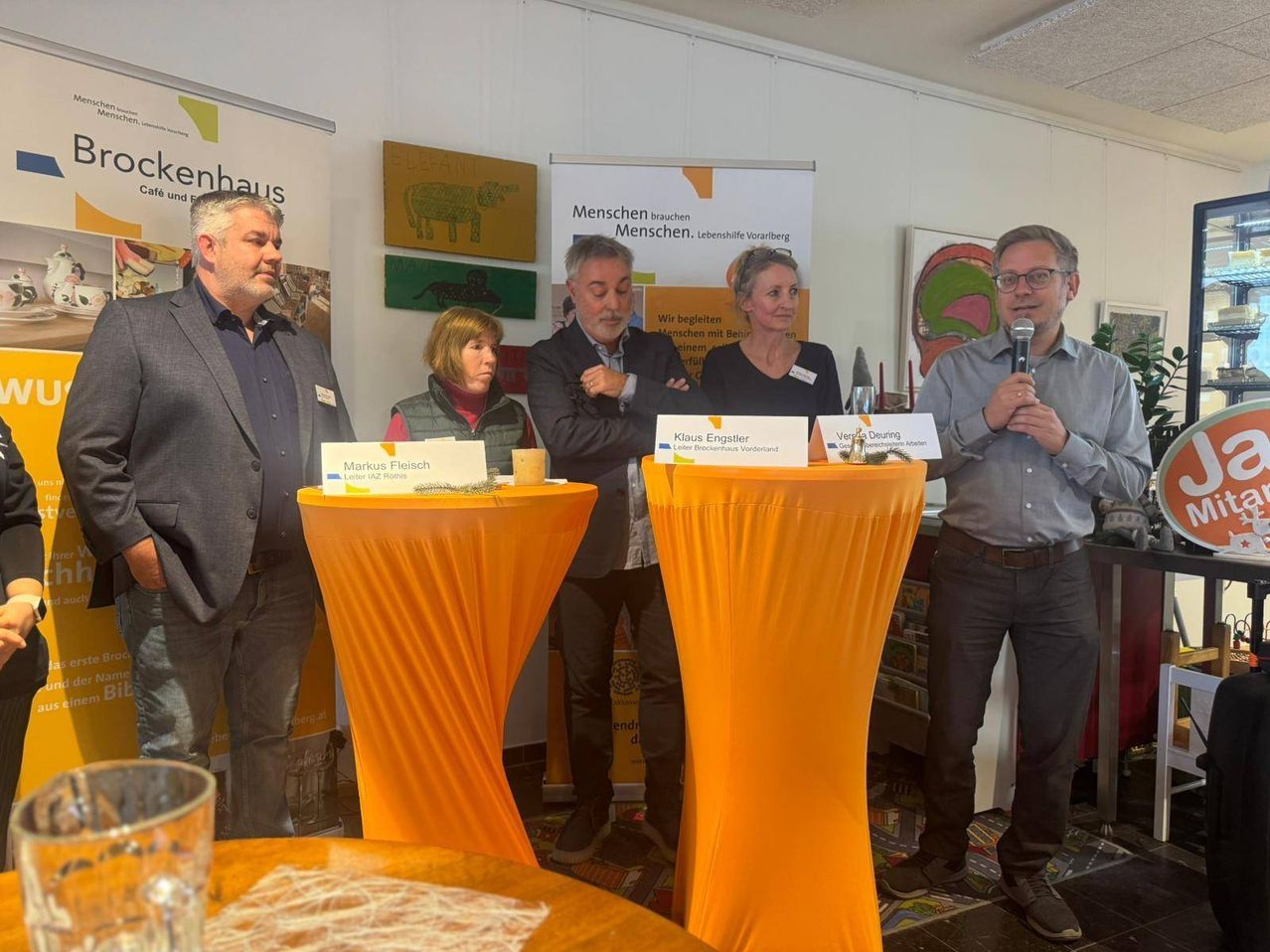 Fünf Personen stehen hinter einem Tisch mit orangefarbenen Tischdecken und blicken in die Menge. Zwei der Personen halten Mikrofone und scheinen zu sprechen. Dahinter steht ein Schild mit der Aufschrift 'Menschen Menschen.' An der Wand befinden sich auch Poster und Gemälde.