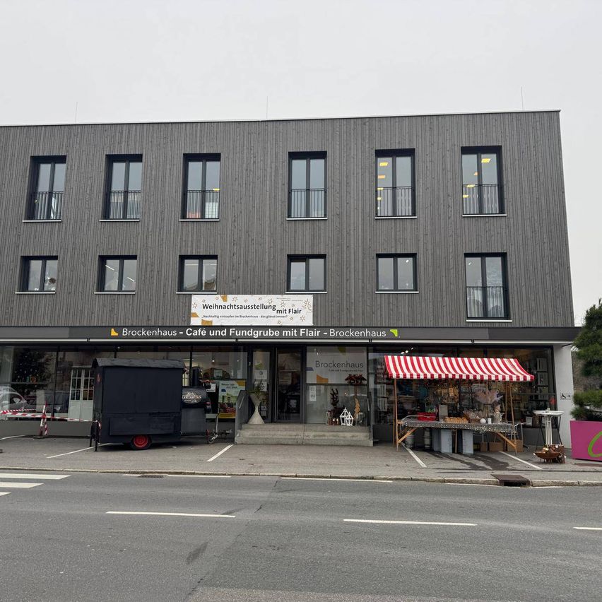 Das Schaufenster von Brockenhaus, einem Café und Spendenzentrum, zeigt einen Weihnachtsmarktstand mit einem rot-weißen Vordach, einem geparkten Imbisswagen und einem klaren Himmel.