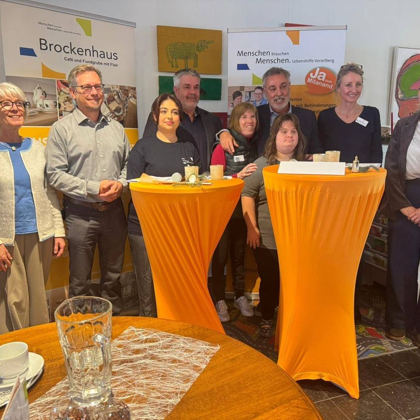 Eine Gruppe von Menschen steht vor einem gelben Tisch mit Tassen und lächelt für ein Foto. Hinter ihnen befinden sich Plakate mit den Texten 'Brockenhaus Cafe und Fundgrube mit Flair' und 'Ja Mitanand'.
