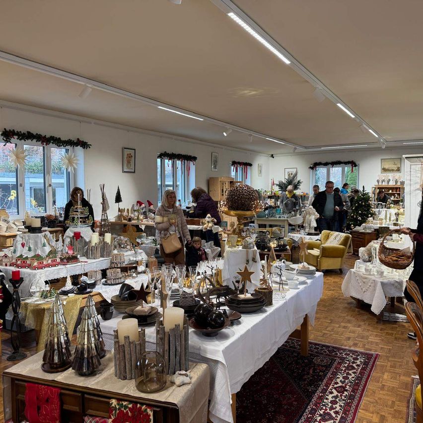 Ein festlicher Markt voller Weihnachtsdekorationen, mit verschiedenen Tischen, auf denen Ornamente, Kerzen und Geschenke stehen. Leute gehen herum und einige kaufen ein. Der Raum ist mit Weihnachtskränzen und Lichtern geschmückt.