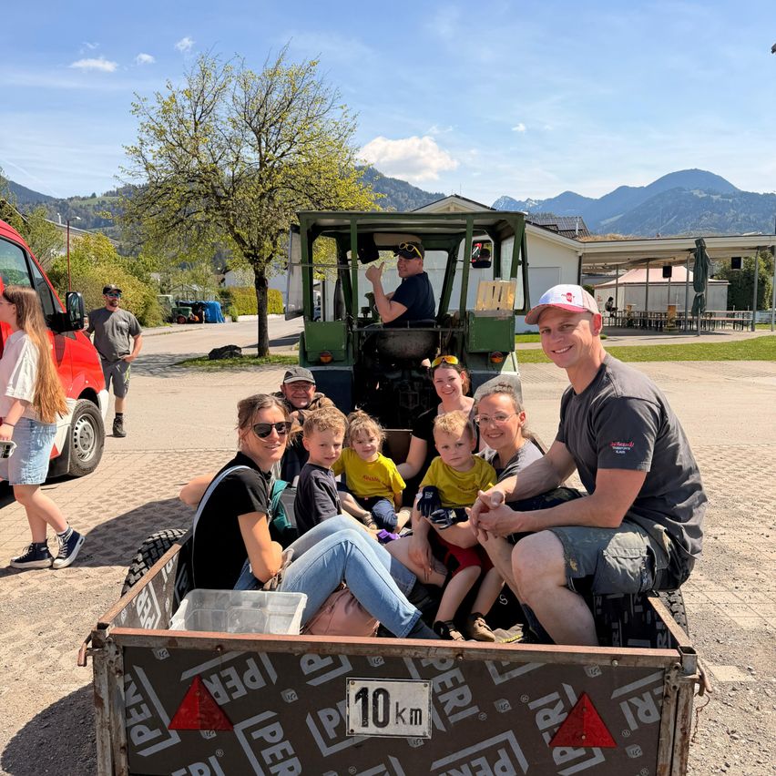 Eine Familie mit sieben Personen sitzt im hinteren Teil eines Traktors. Das Fahrzeug ist auf der Straße geparkt, und im Hintergrund ist ein Gebäude mit einem Baum und Bergen zu sehen.