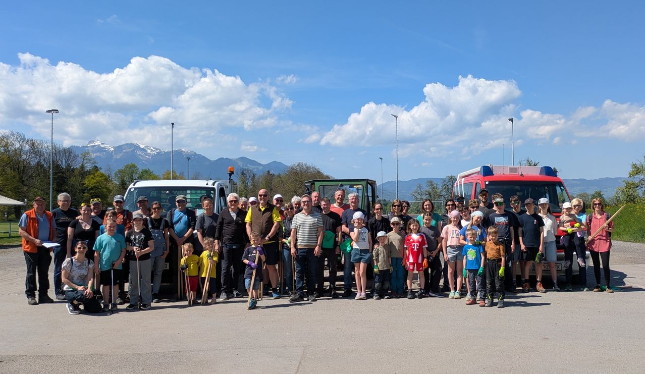 Eine Gruppe von Menschen steht vor einem weißen und grünen Lkw mit Bergen im Hintergrund. Sie halten Stöcke und tragen Hüte und Sonnenbrillen.