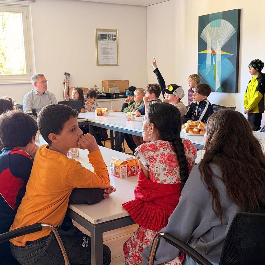 Kinder sitzen an Tischen und hören einem Erwachsenen in einem Klassenzimmer zu. Ein Gemälde hängt an der Wand.