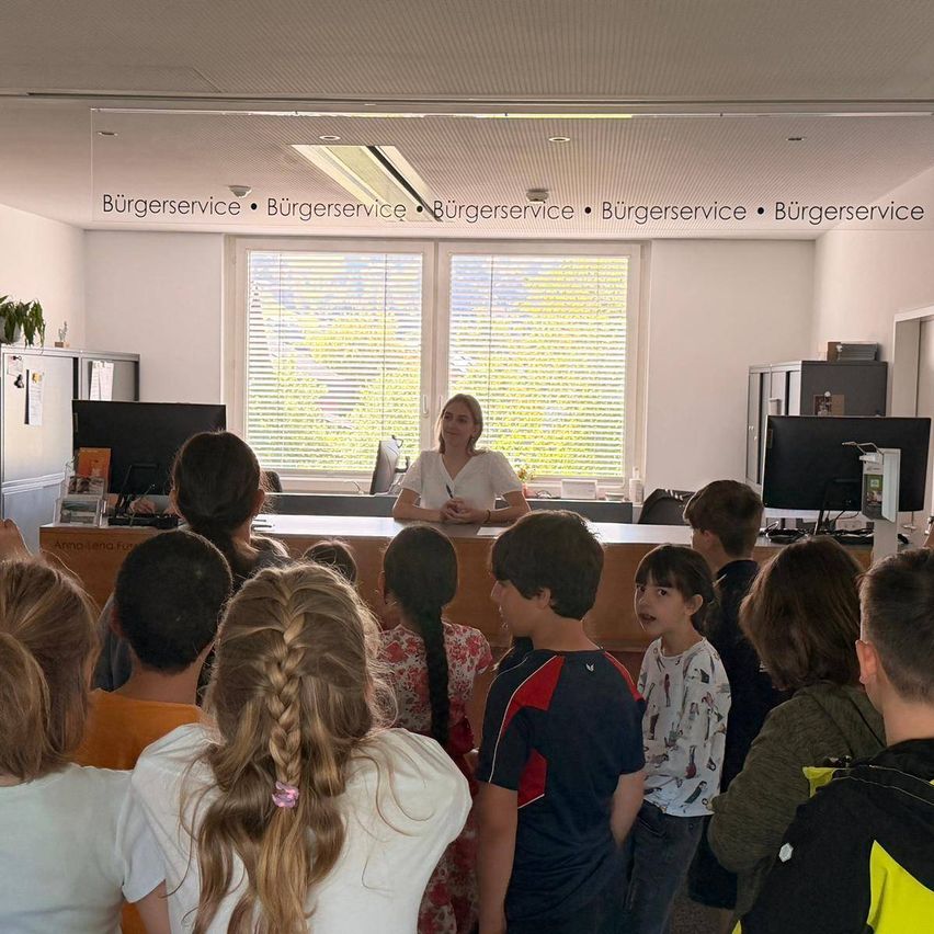 Eine Frau steht hinter einer Theke in einem Raum mit Kindern. Sie steht vor einer Gruppe von Kindern, die aufmerksam zuhören. Der Raum hat ein Fenster mit Jalousien und ein Schild mit dem Wort Burgerservice.