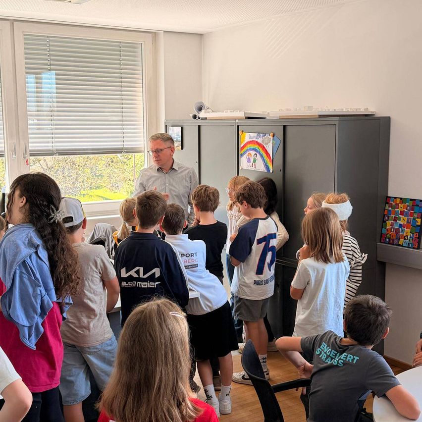 Ein Mann steht vor einer Gruppe von Kindern in einem Raum mit weißer Wand und Holzboden. Er spricht mit ihnen.