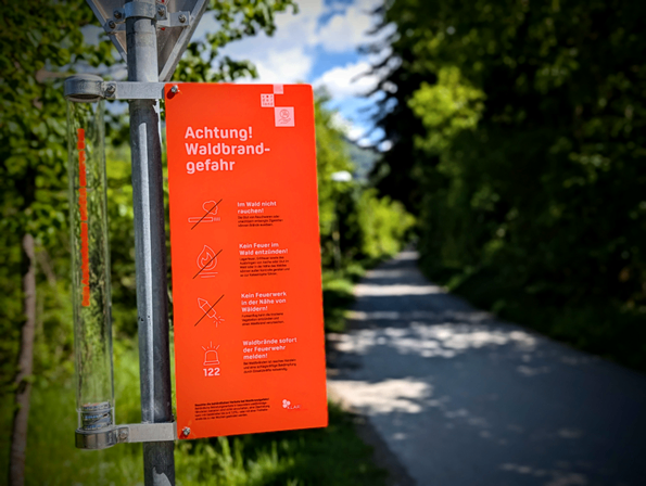 Ein rotes Schild an einem Pfosten warnt vor 'Achtung! Waldbrandgefahr' mit Anweisungen, im Wald nicht zu rauchen und keine Feuer zu entfachen. Ein Weg führt durch den Wald.