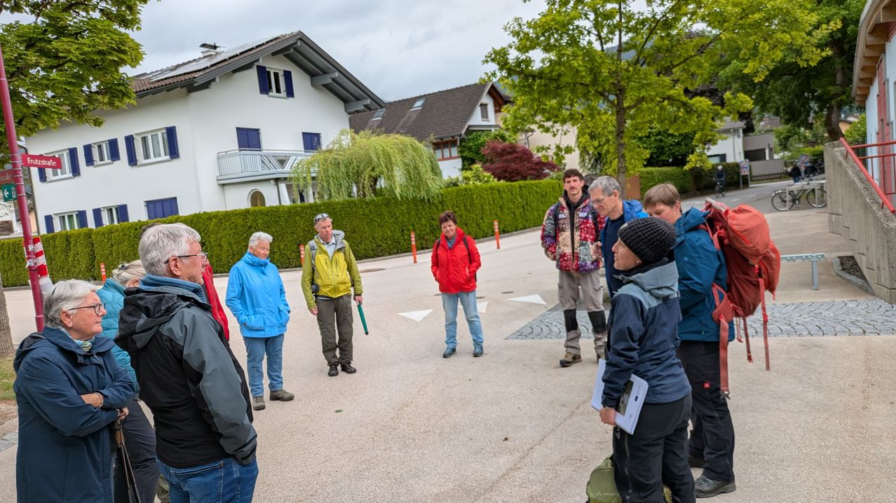 Eine Gruppe von Menschen, die für das Wetter gekleidet sind, steht auf einem Parkplatz, möglicherweise zu Beginn einer Tour. Bäume und Häuser sind im Hintergrund zu sehen.