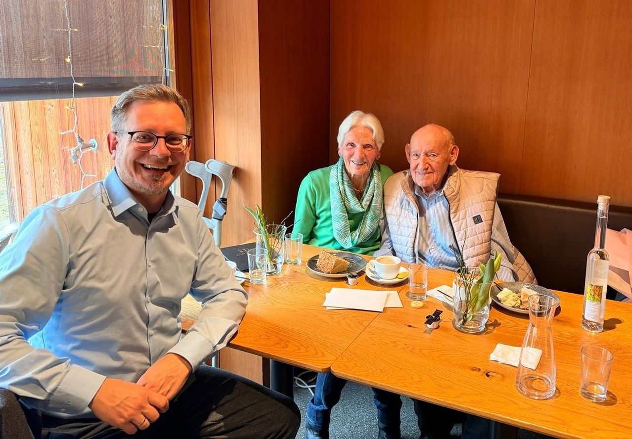 Drei Personen sitzen um einen Holztisch herum und lächeln für ein Foto. Auf dem Tisch stehen ein Teller, eine Tasse, eine Untertasse und eine Vase mit Blumen. Der Mann trägt eine Brille und einen Ring. Die Frau trägt einen Schal. Der ältere Mann trägt eine Weste.