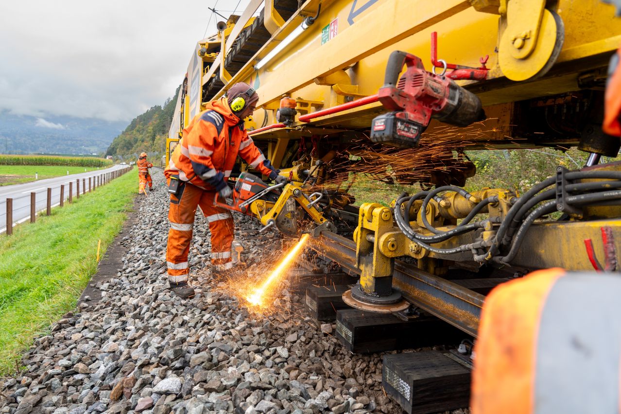 A worker in safety gear uses a grinder to cut through railroad tracks, with sparks flying, near a yellow maintenance vehicle.